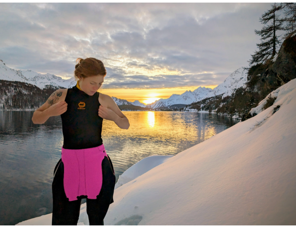A person in a black and pink wetsuit stands beside a serene lake at sunset, adjusting their suit while surrounded by snow-covered mountains and trees.